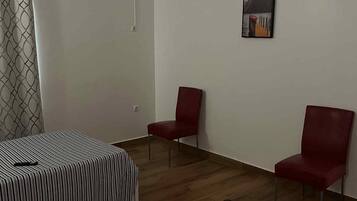 Une pièce avec un lit, deux chaises rouges, une couverture à carreaux, un tableau et une fenêtre avec un rideau.