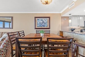 Uma área de refeições com uma mesa de madeira, cadeiras, uma obra de arte emoldurada e uma cozinha ao fundo.