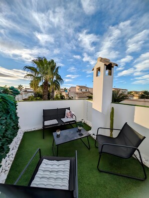 An outdoor patio with a sofa, chairs, and a table, surrounded by greenery and a clear sky.