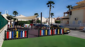 Una calle soleada con un parque infantil, coches aparcados y edificios residenciales.