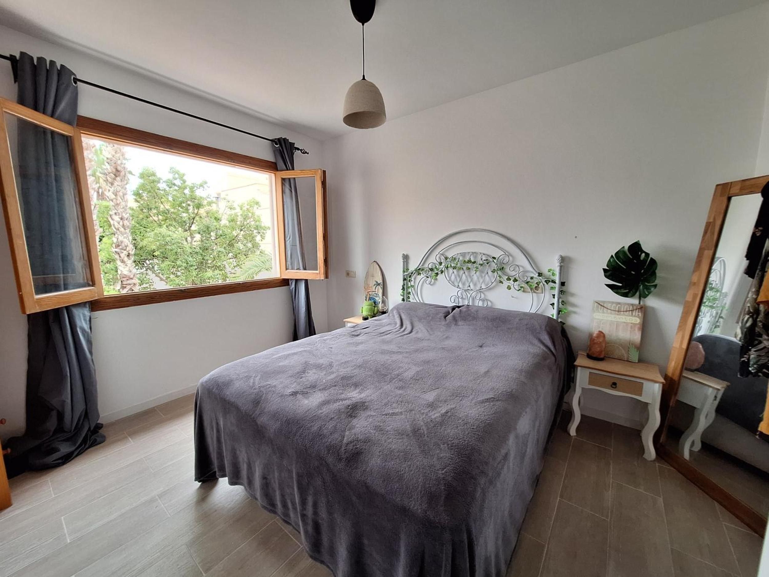A bedroom with a bed, a window with curtains, a bedside table, a nightstand, a mirror, and a hanging light fixture.