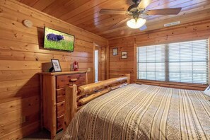 Quarto de cabana de madeira com cama, ventoinha no teto, televisão montada e janela com persianas.