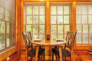 Un intérieur en bois avec une table ronde et quatre chaises, de grandes fenêtres équipées de stores et une vue sur les arbres à l’extérieur.