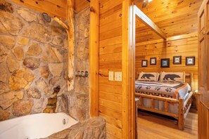 L’intérieur d’une cabane en rondins, avec un mur en pierre, un lit en bois agrémenté de linge de lit à motifs d’ours et une baignoire blanche.