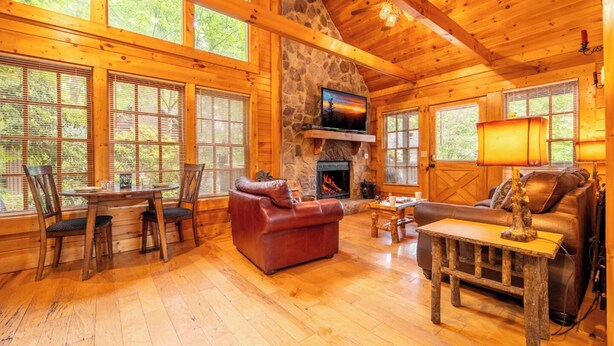 A cozy cabin interior with a stone fireplace, wooden furniture, and large windows. - Serene 1BR Cabin - Hot tub & Fireplace - Smokies (Sevierville)