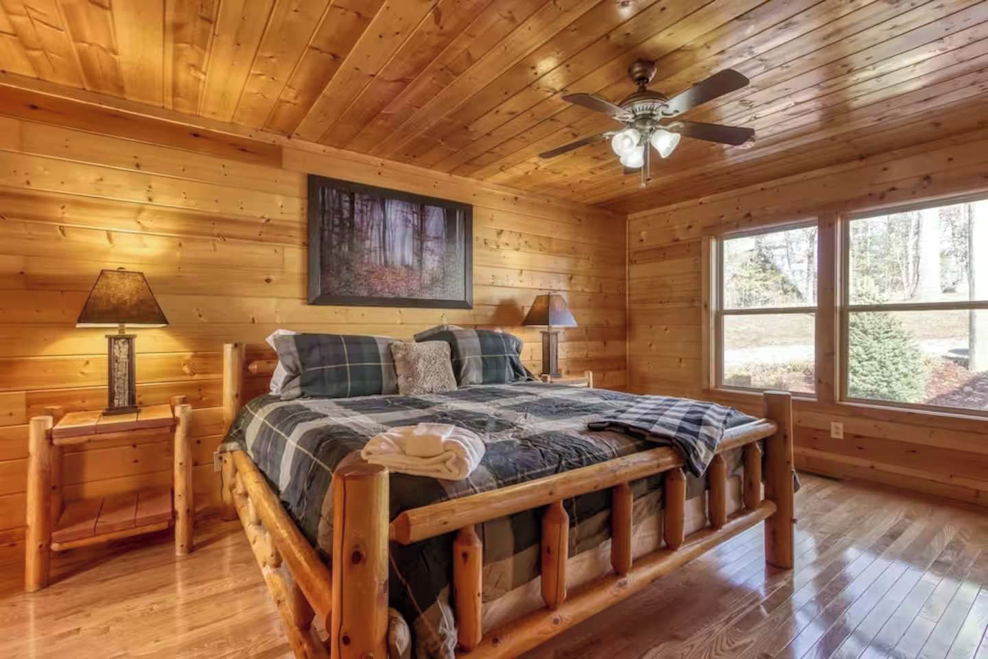 A wooden cabin bedroom with a bed, two bedside lamps, a ceiling fan, and a large window.