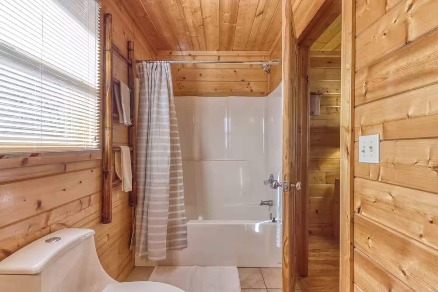 A wooden cabin bathroom with a shower, toilet, and a window with blinds.