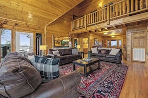 A spacious log cabin interior with a high ceiling, a balcony, and a fireplace. - Bold 7BR Cabin - Home Theater & Hot Tub - Smokies (Sevierville)