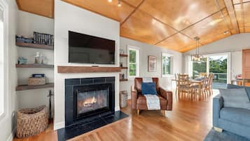A spacious living room with a fireplace, wooden flooring, a leather armchair, and a dining area with natural light.