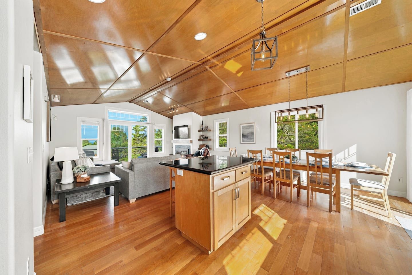 A modern kitchen with wooden cabinetry, a dining area with a wooden table and chairs, and a large window providing a view of greenery outside.
