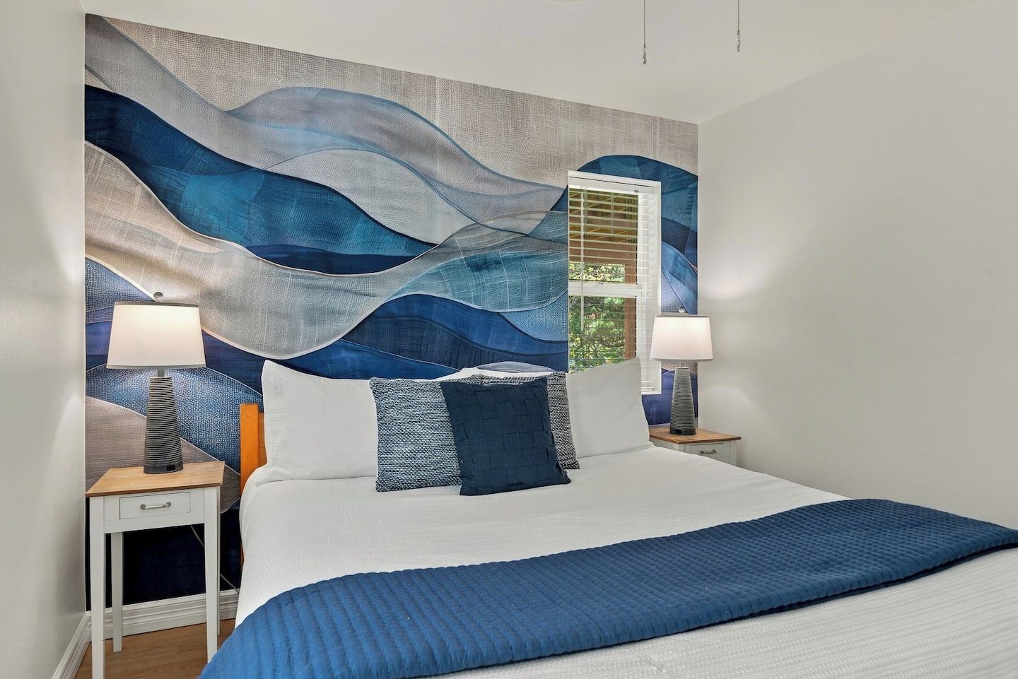 A bedroom with a large bed, two bedside tables, a wall with a textured abstract design, and a window with blinds.