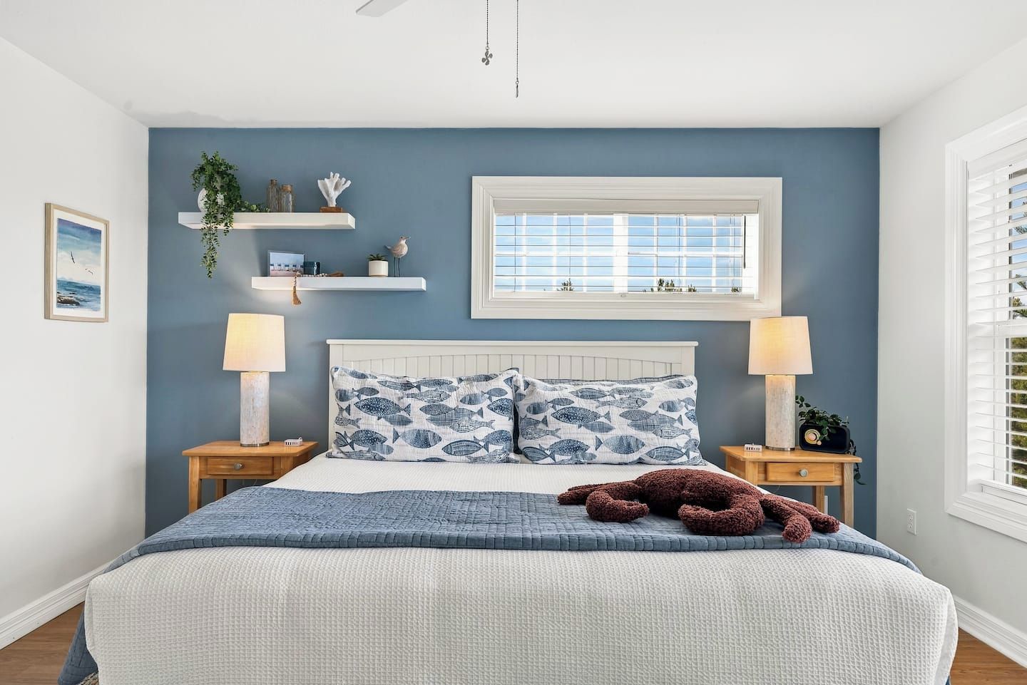 A bedroom with a bed, two bedside tables, a window with blinds, a wall-mounted shelf with decorative items, and a hanging plant.