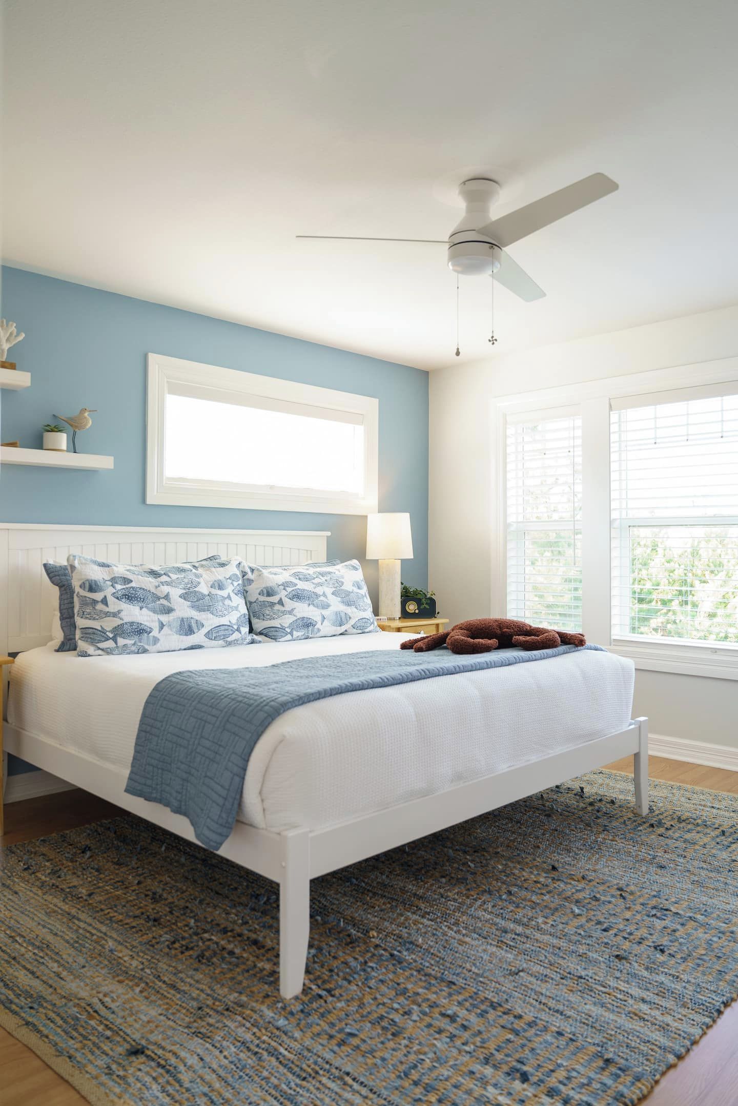 A bedroom with a bed, a ceiling fan, a window with blinds, and a nightstand with a lamp.