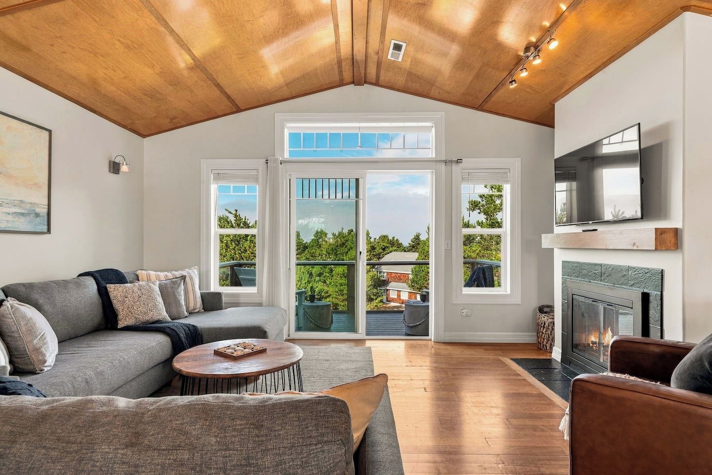 A spacious living room with a high ceiling, a fireplace, a television mounted above it, and a large sliding glass door leading to an outdoor deck.