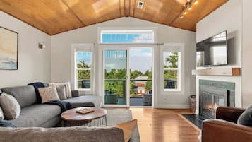 A spacious living room with a high ceiling, a fireplace, a television mounted above it, and a large sliding glass door leading to an outdoor deck.