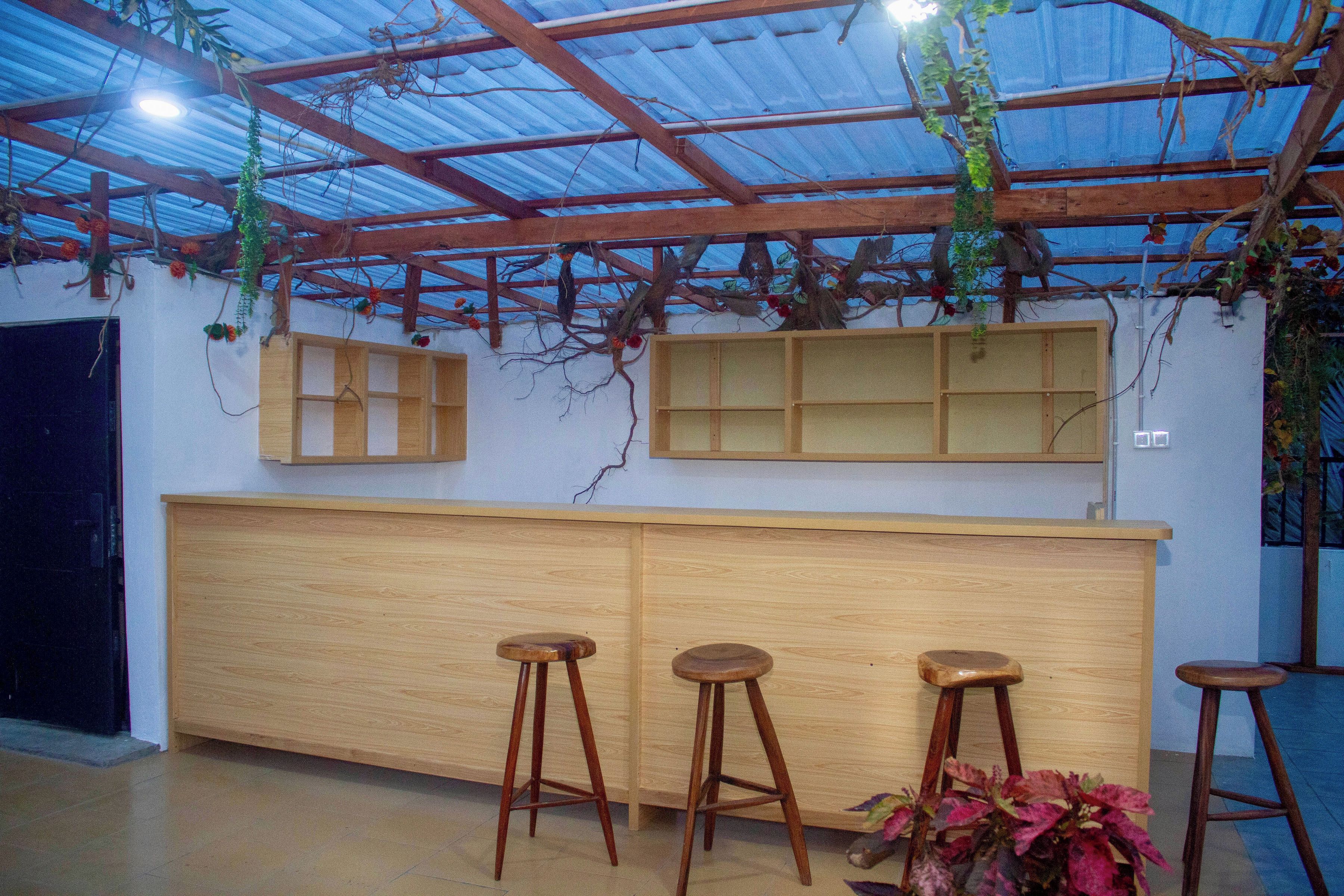 Zona de bar con taburetes de madera y una barra, bajo un techo metálico con plantas.