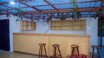 Zona de bar con taburetes de madera y una barra, bajo un techo metálico con plantas.