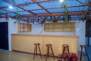 A bar area with wooden stools and a counter, under a metal roof with plants.
