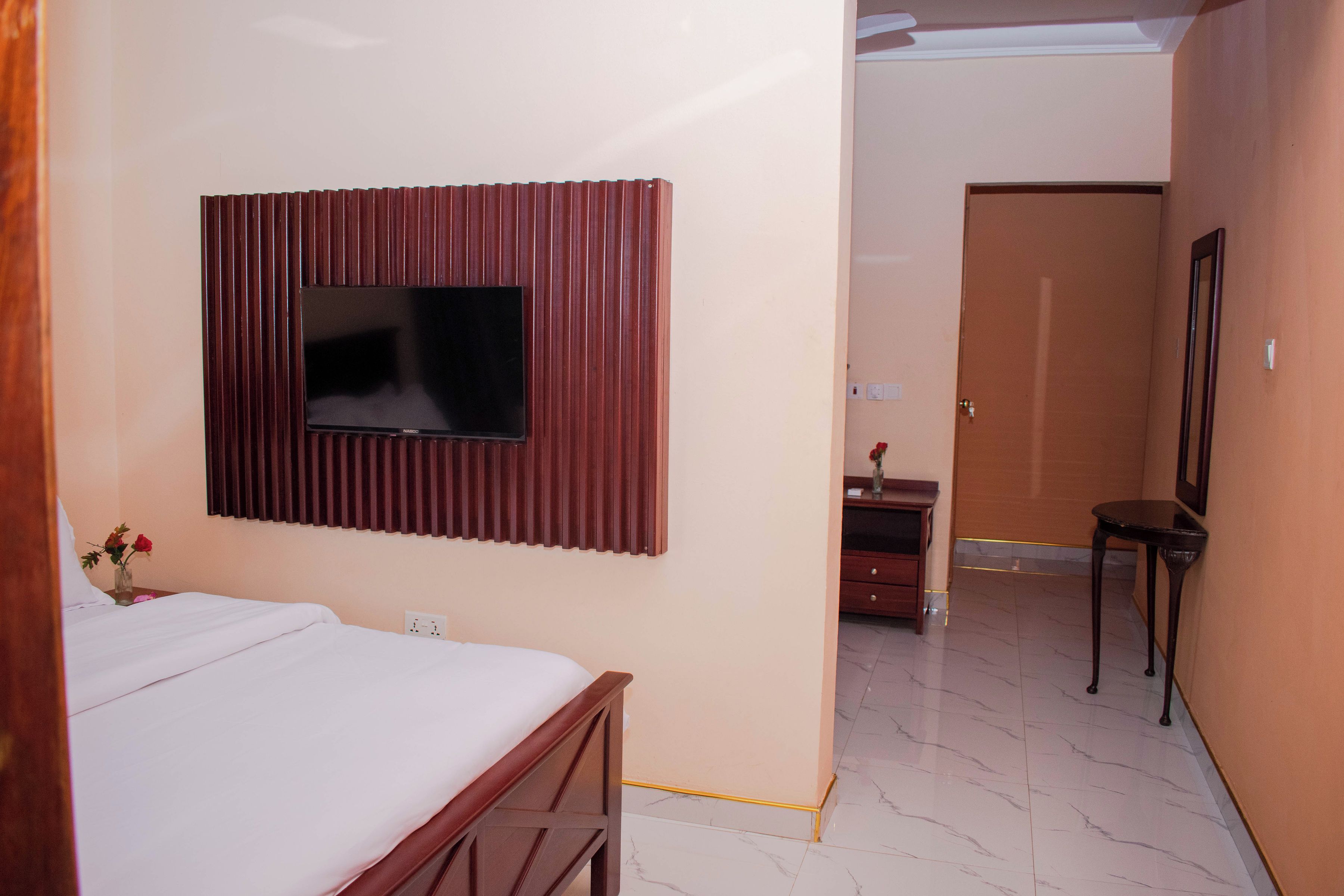 Una habitación de hotel con cama, televisor montado en la pared, un mueble de madera y una puerta que da a otra habitación.