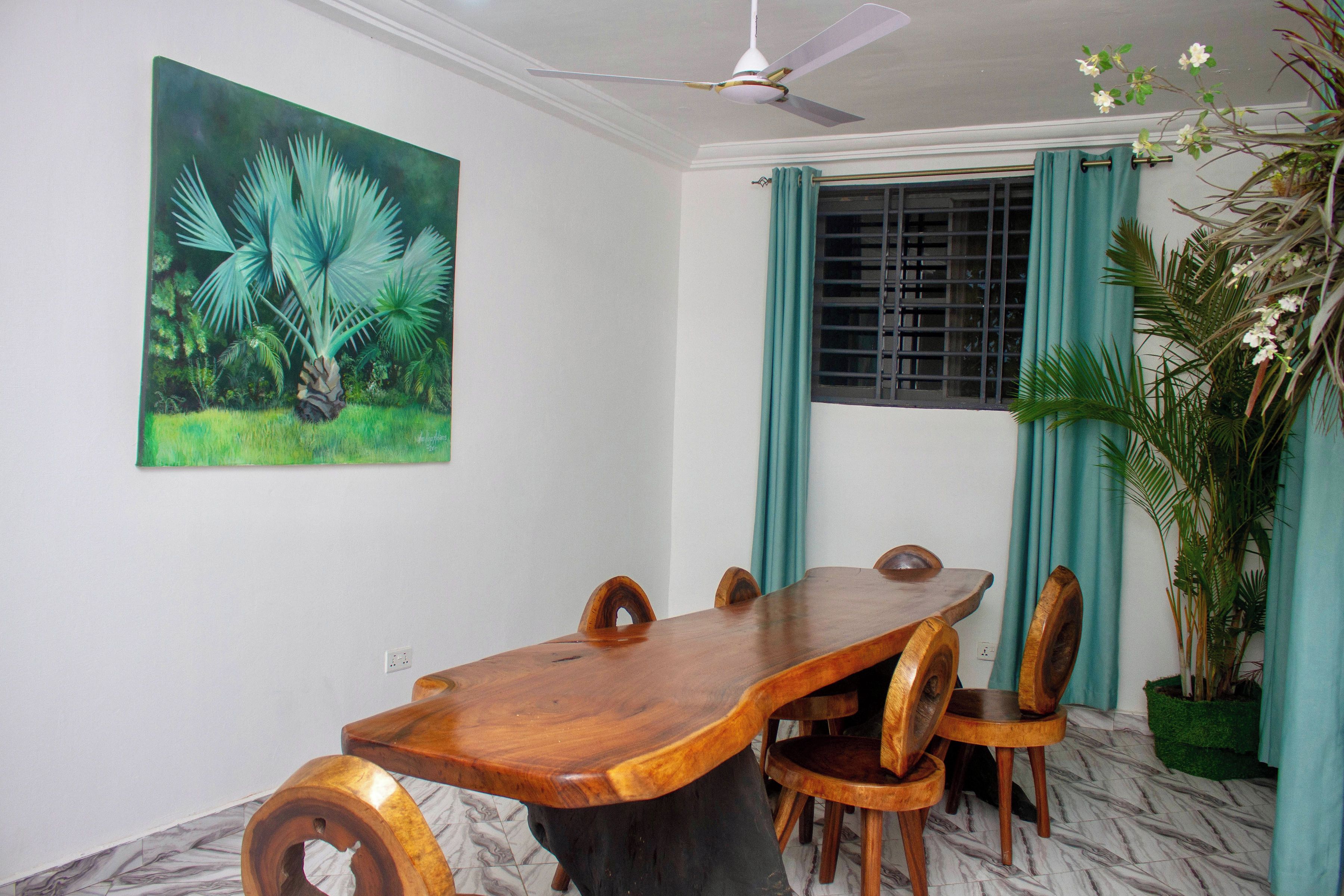 Uma área de refeições com mesa e cadeiras de madeira, ventoinha no teto, janela com cortinas e um quadro com folhagem tropical na parede.