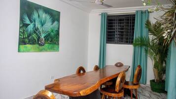 Uma área de refeições com mesa e cadeiras de madeira, ventoinha no teto, janela com cortinas e um quadro com folhagem tropical na parede.