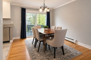A dining area with a wooden table, six chairs, a chandelier, and a view through a large window. - Fully Renovated 4BR Home, 2 Baths, Deck, Parking (Saratoga Springs)