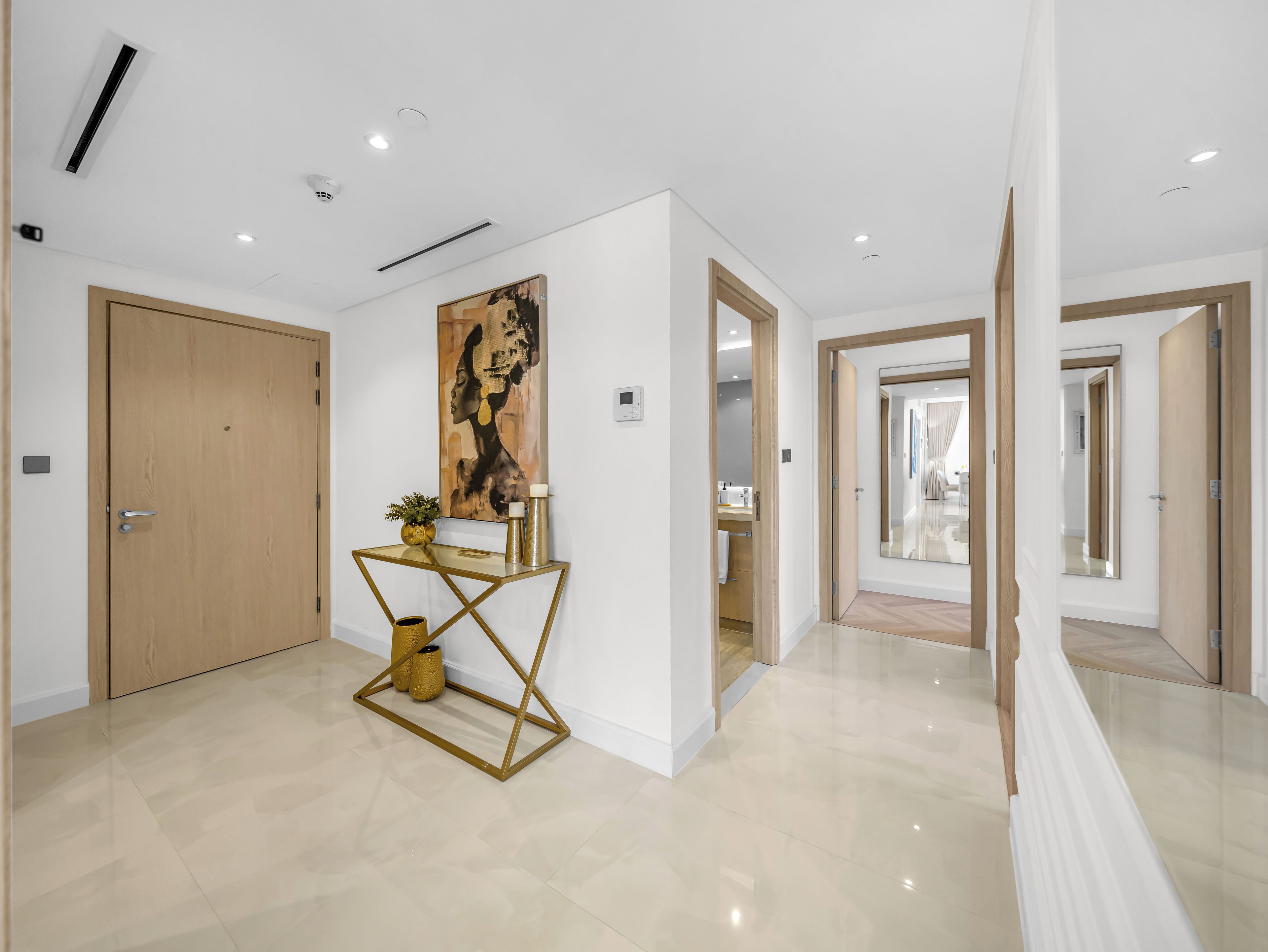 Un interior moderno con una puerta de madera, una consola de vidrio con objetos decorativos y un gran cuadro abstracto en la pared.