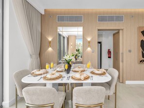 A dining area with a white table set for four, featuring gold-toned plates and cutlery, a vase with white flowers, and a city view through large windows. - Kensington Iconic Burj View Dubai Mall (Dubai)