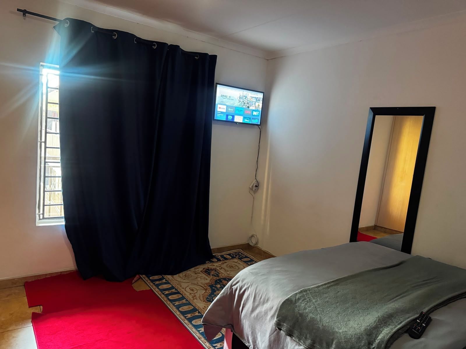 A bedroom with a bed, a television mounted on the wall, a window with curtains, and a mirror.