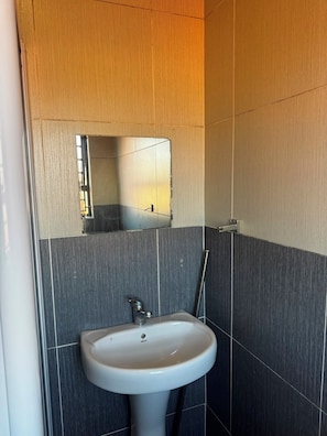 A bathroom with a white pedestal sink, a mirror, and dark grey tiled walls. - Room-charming Apartment Room in Johannes (Johannesburg)