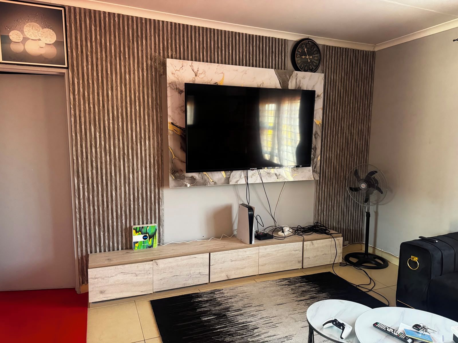 A modern living room with a flat-screen TV mounted on a marble wall, a wooden entertainment unit below, a black and white rug, and a black suitcase.