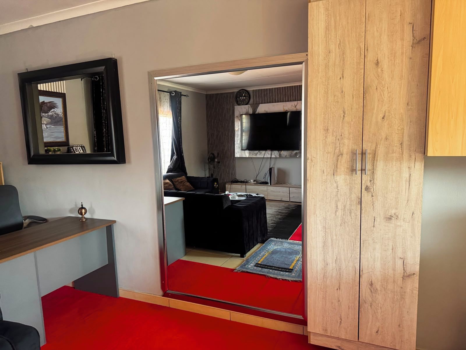 A room with a large mirror, a wooden wardrobe, a red carpet, a black sofa, and a television in the background.