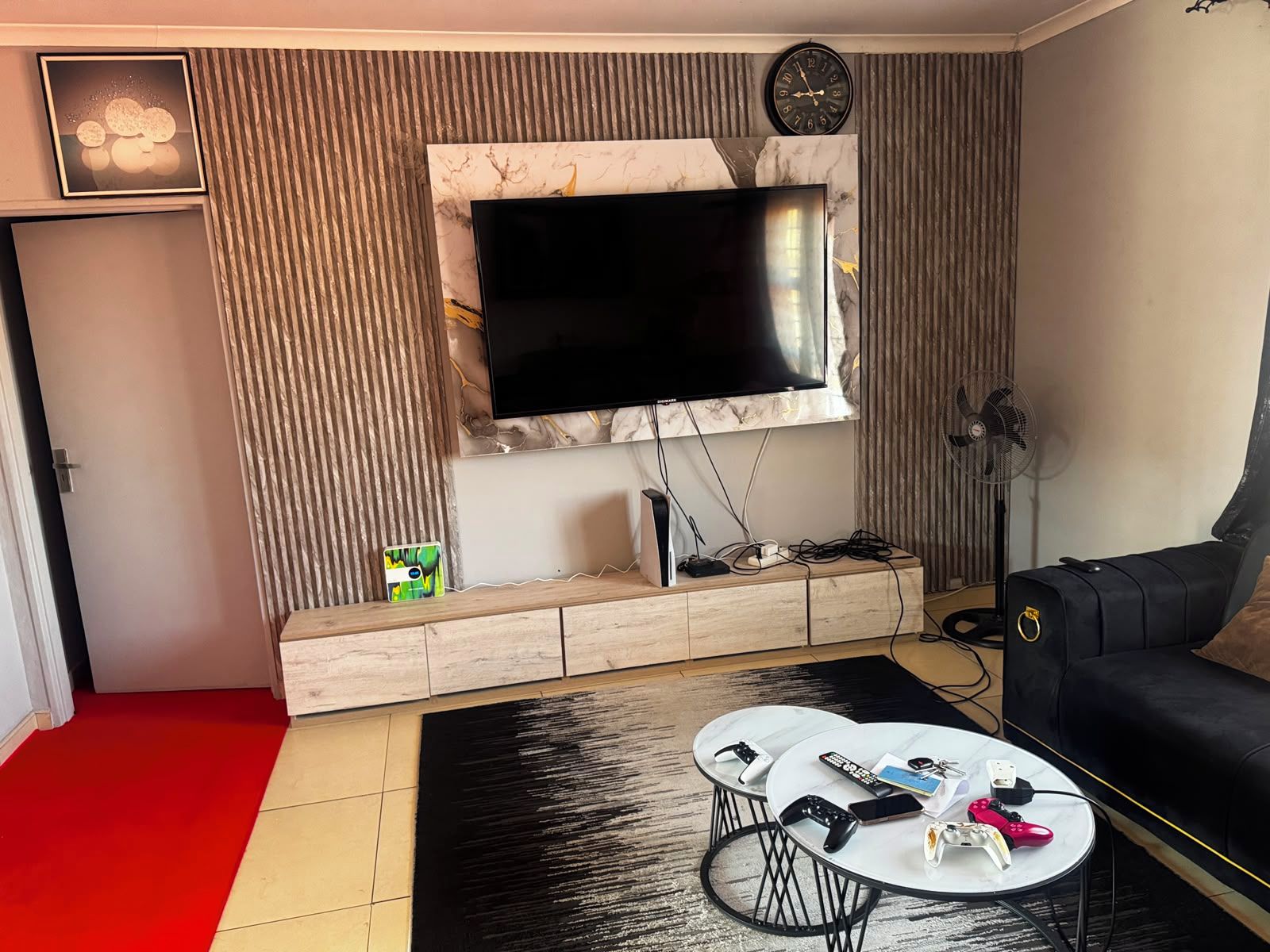 A modern living room with a flat-screen TV mounted on a marble wall, a wooden entertainment unit, a black sofa, and a coffee table with gaming controllers.
