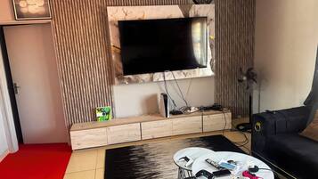 A modern living room with a flat-screen TV mounted on a marble wall, a wooden entertainment unit, a black sofa, and a coffee table with gaming controllers.