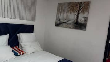 A bedroom with a bed, white bedding, a blue headboard, a painting of a tree, and a window with blinds.