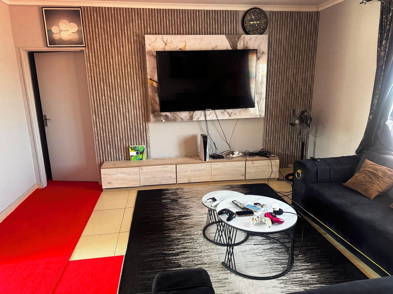 A modern living room with a flat-screen TV mounted on a marble wall, a black sofa with a brown cushion, a coffee table with remotes and a game controller, and a red and beige carpet.