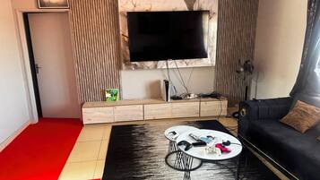 A modern living room with a flat-screen TV mounted on a marble wall, a black sofa with a brown cushion, a coffee table with remotes and a game controller, and a red and beige carpet.