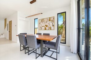 A modern dining room with a wooden table and grey upholstered chairs, a large abstract painting on the wall, and a view of palm trees through a glass door. - 25% Off Oct 7-15, Private Pool, Walk 2 Beach/Shops (Grace Bay)