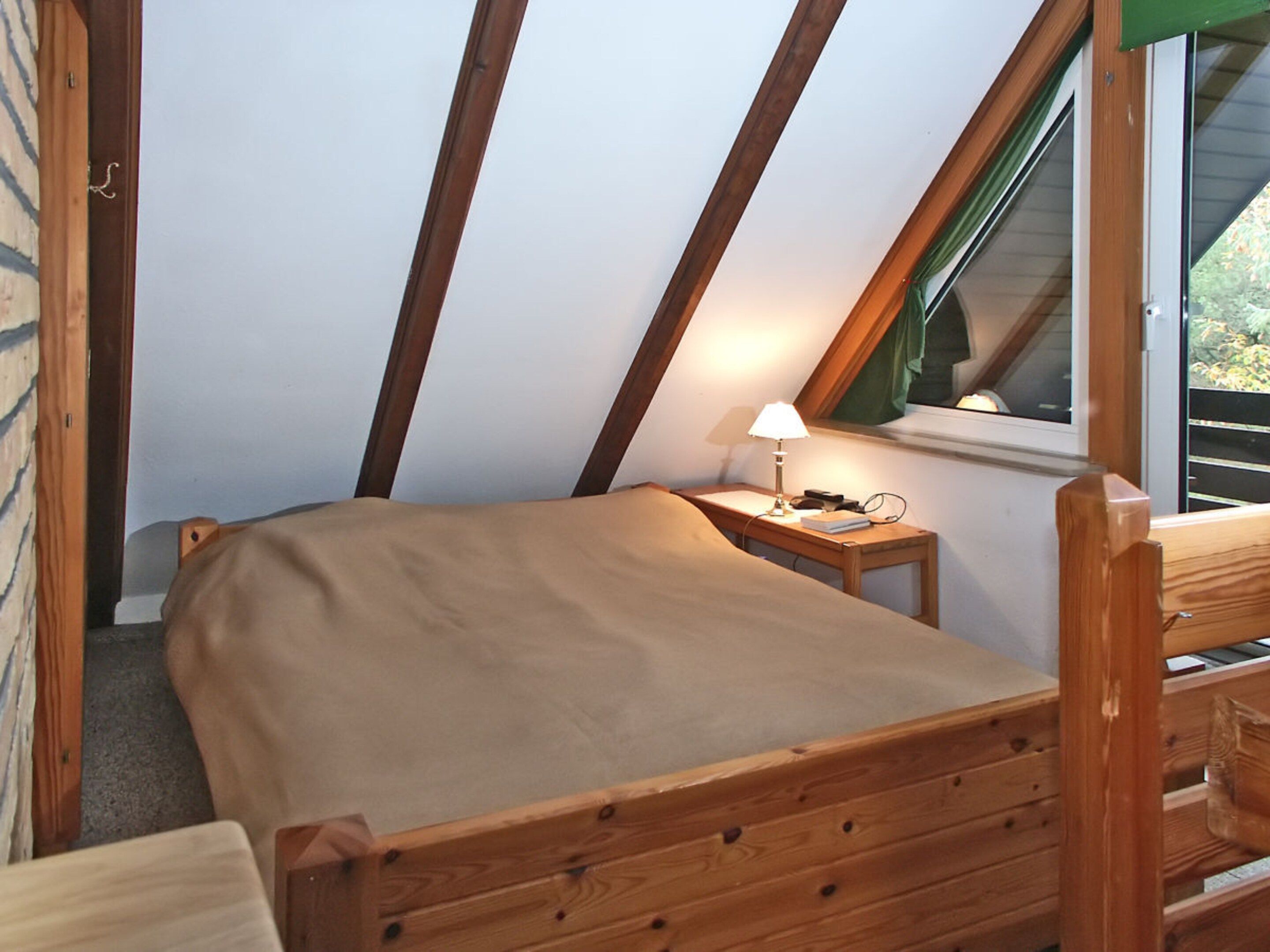 Um quarto pequeno e acolhedor, com teto inclinado, uma cama de solteiro com cabeceira de madeira, uma mesinha de cabeceira com abajur e uma janela com vista para o verde.