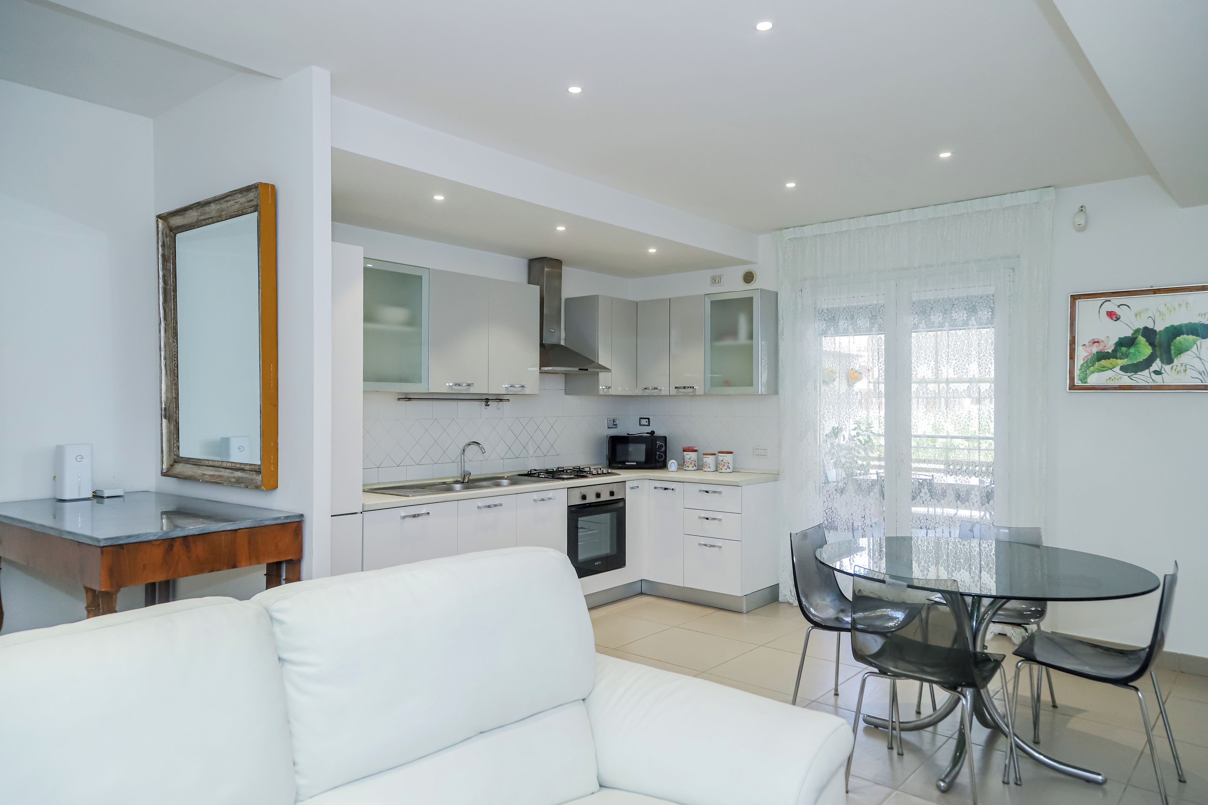 A modern kitchen with white cabinets, a stove, and a microwave. There is a dining area with a glass table and chairs.
