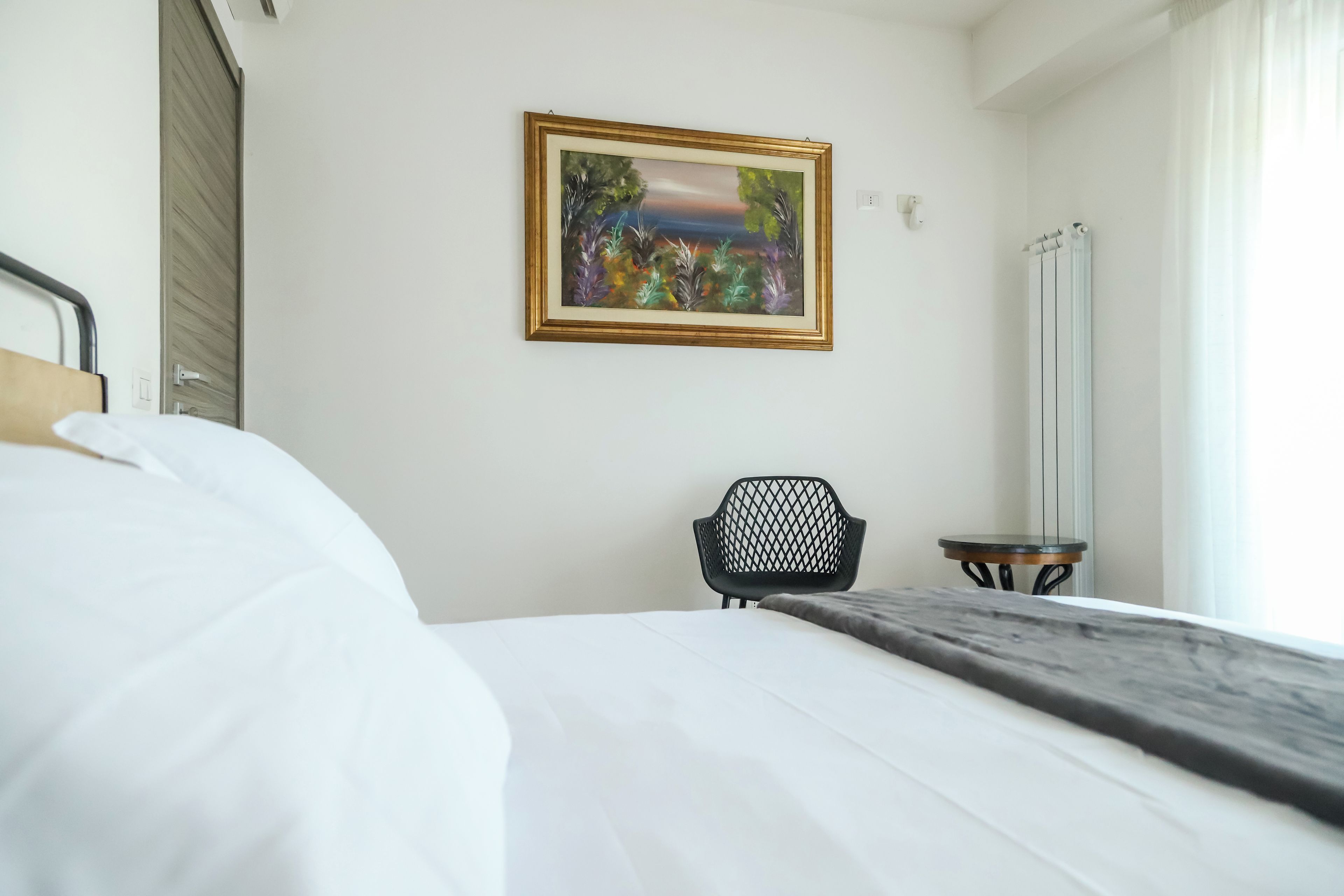 A hotel room with a bed, a chair, a painting on the wall, and a window with curtains.
