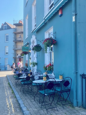 An outdoor seating area with tables and chairs, a building with blue walls, and hanging flower baskets.