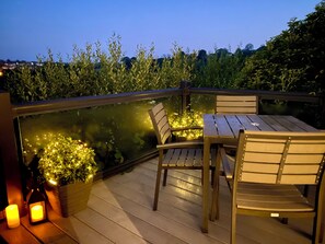 Une terrasse en bois avec une table et des chaises, entourée de verdure et protégée par une barrière en verre.