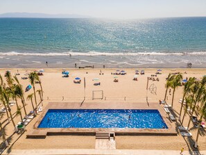 A poolside view of a beach resort with a swimming pool, beach umbrellas, and palm trees. - Aria 3BR Private Jacuzzi & Terrace Nuevo Vallarta (Nuevo Vallarta)