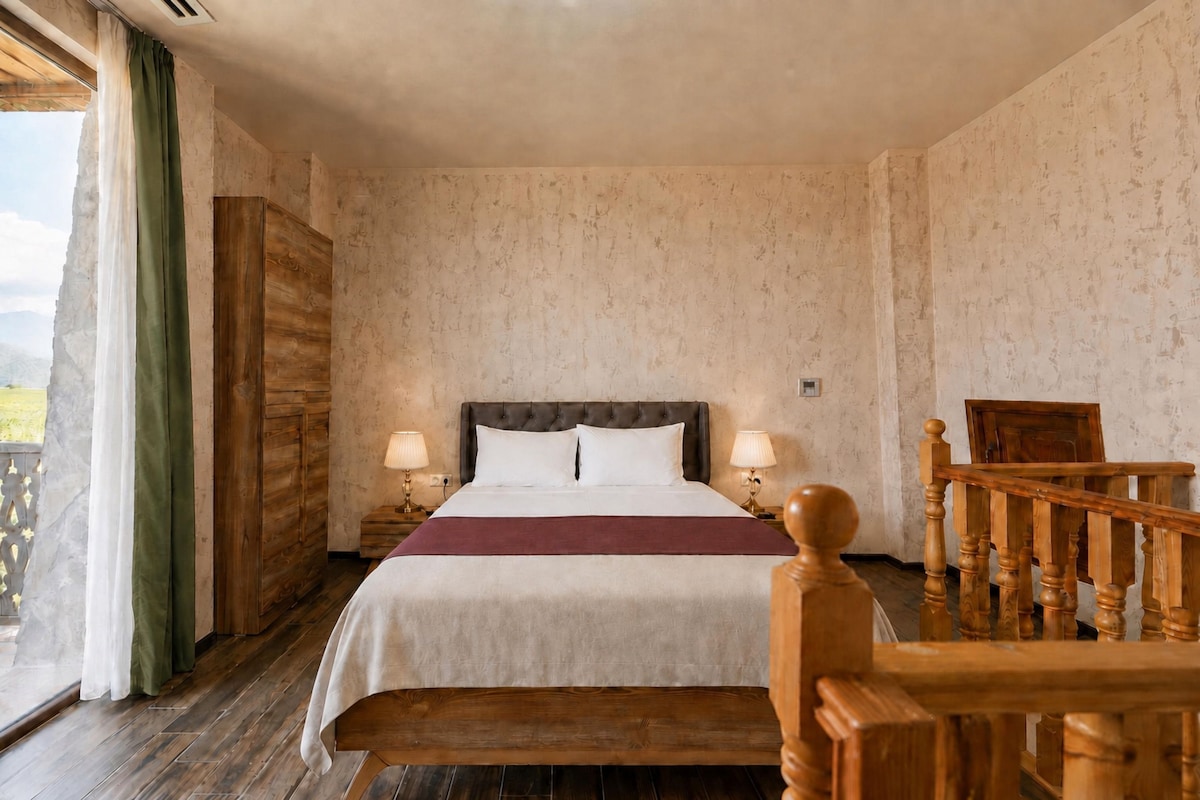 A hotel room with a bed, two bedside lamps, a wooden headboard, a view of the outdoors, and a wooden staircase.