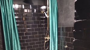 Un baño con paredes de azulejos negros, una ducha con rociador dorado y un lavamanos negro.