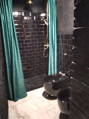 Ein Badezimmer mit schwarzen Fliesenwänden, einer Dusche mit goldfarbener Brause und einem schwarzen Waschbecken.