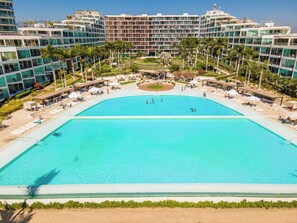 A large, clear blue pool surrounded by lounge chairs and umbrellas, with modern buildings in the background. - Aria!! New 2 Bedroom with Gorgeous Pool and views (Nuevo Vallarta)