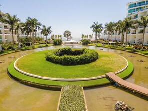 A circular water feature with a central island, surrounded by a pool, palm trees, and a building with balconies. - Aria!! New 2 Bedroom with Gorgeous Pool and views (Nuevo Vallarta)