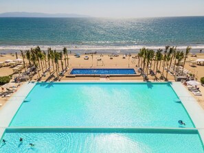 Piscina con vista a la playa y al océano, rodeada de palmeras y tumbonas.